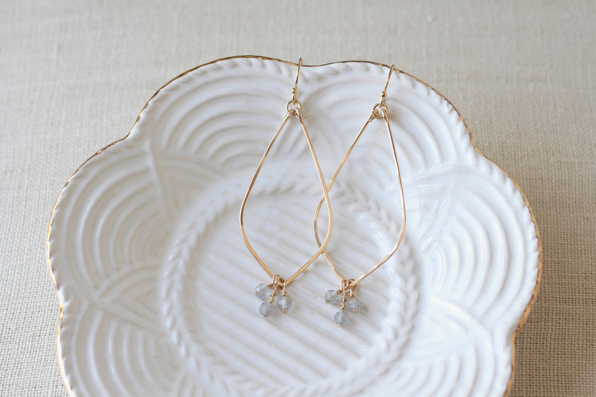 Gold teardrop dangle earrings with bluish gray gemstone clusters, displayed on a white scalloped ceramic dish over a beige fabric background.