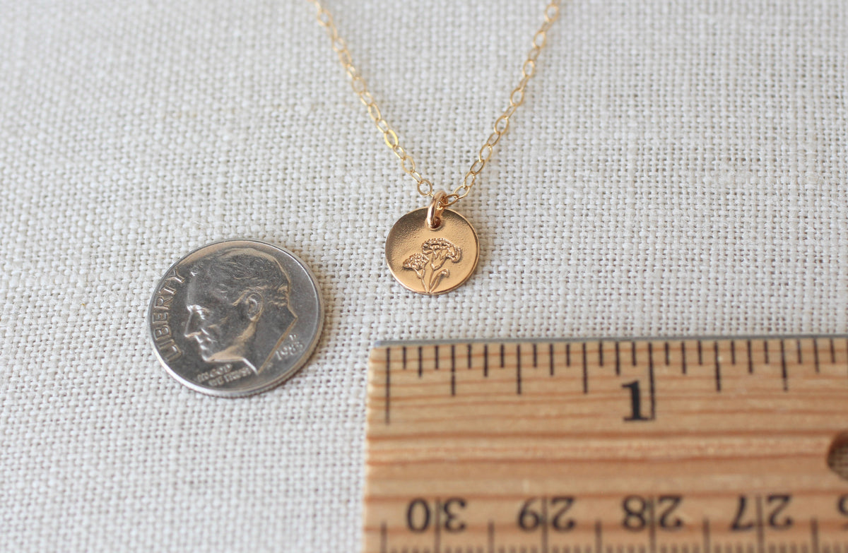 A delicate gold necklace with a small round disc pendant engraved with a flower, displayed on a textured beige fabric alongside a dime for scale and a wooden ruler.