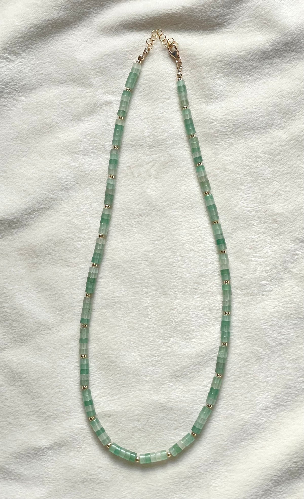 A beaded necklace made of light green cylindrical beads separated by small gold spacer beads, with a gold clasp, laid flat on a white textured fabric background.