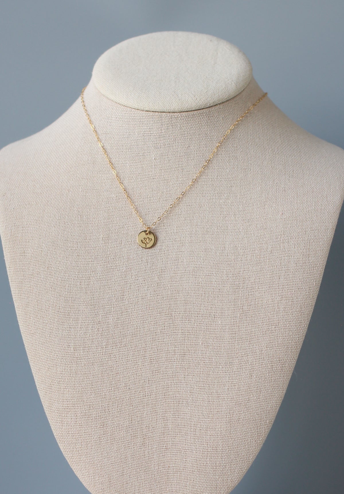 A delicate gold chain necklace with a small circular engraved pendant, displayed on a cream fabric jewelry bust against a soft blue-gray background.