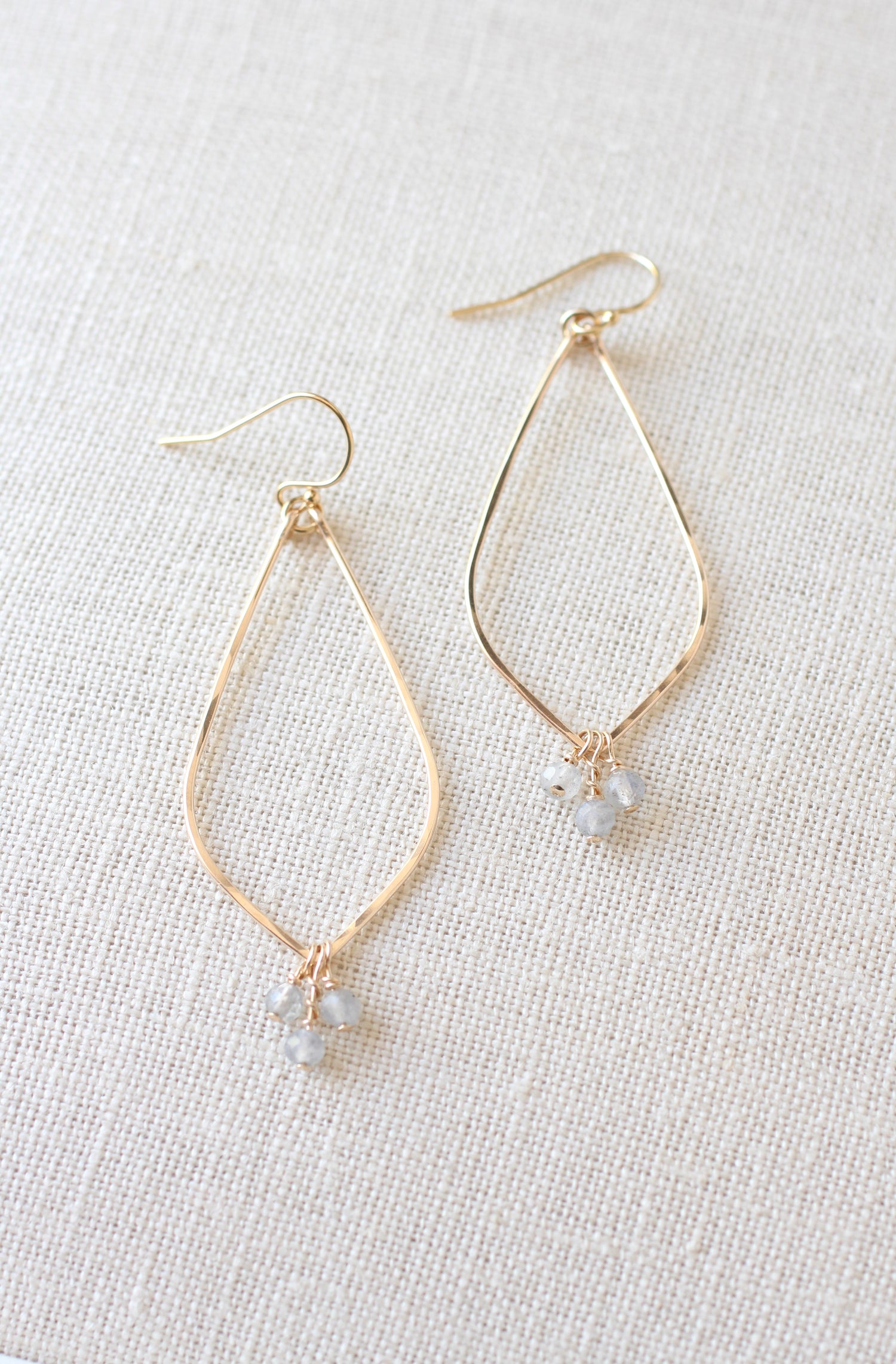 Gold-tone teardrop dangle earrings with small gray faceted beads, displayed on a textured beige linen background.