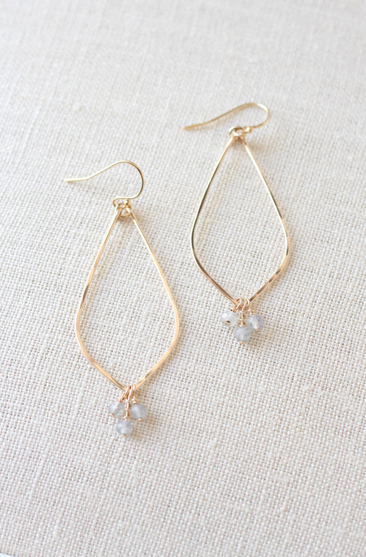 Gold-tone teardrop dangle earrings with small gray faceted beads, displayed on a textured beige linen background.