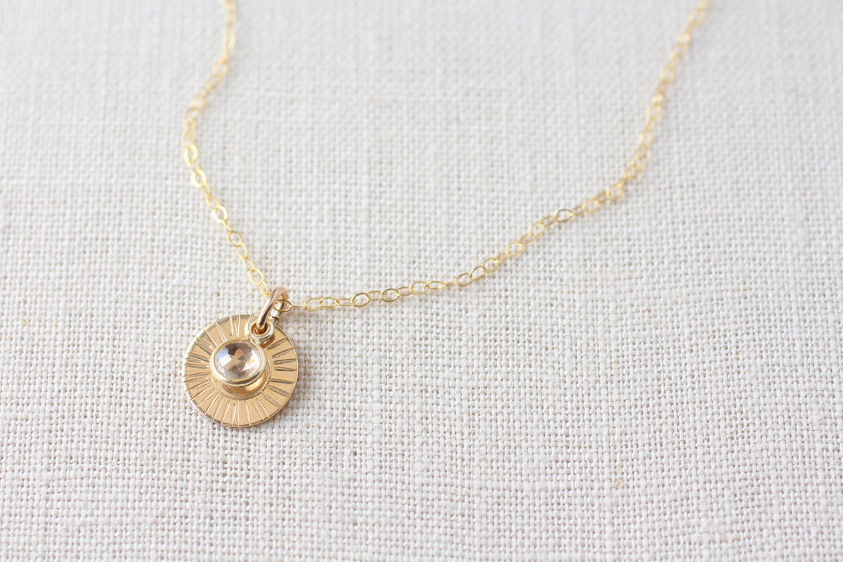 A delicate gold chain necklace featuring a small sunburst disc pendant with a stone center, displayed on a neutral beige linen fabric background.
