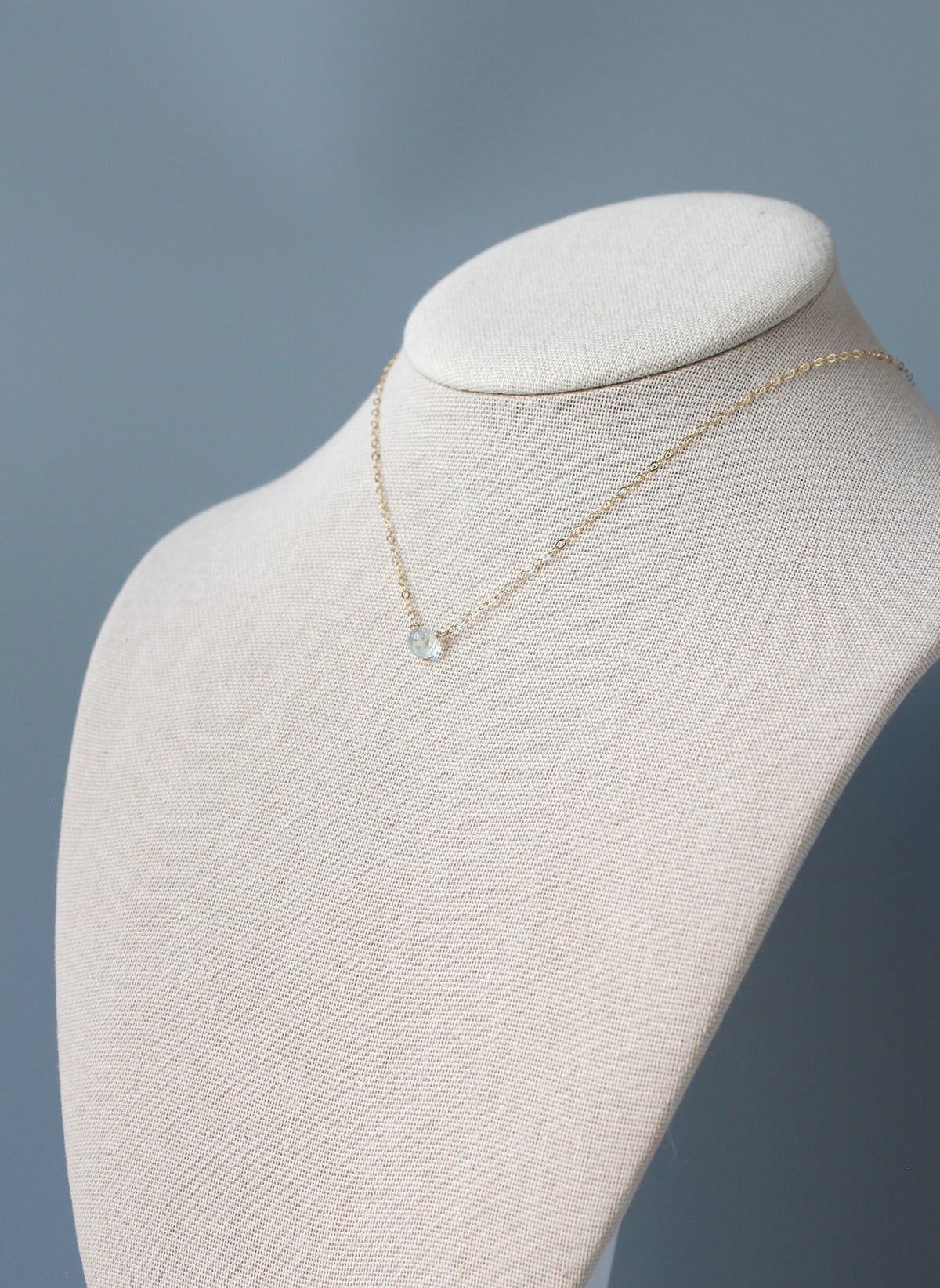 A delicate gold chain necklace with a small light green  faceted stone pendant, displayed on a cream-colored fabric jewelry bust against a soft blue-gray background.