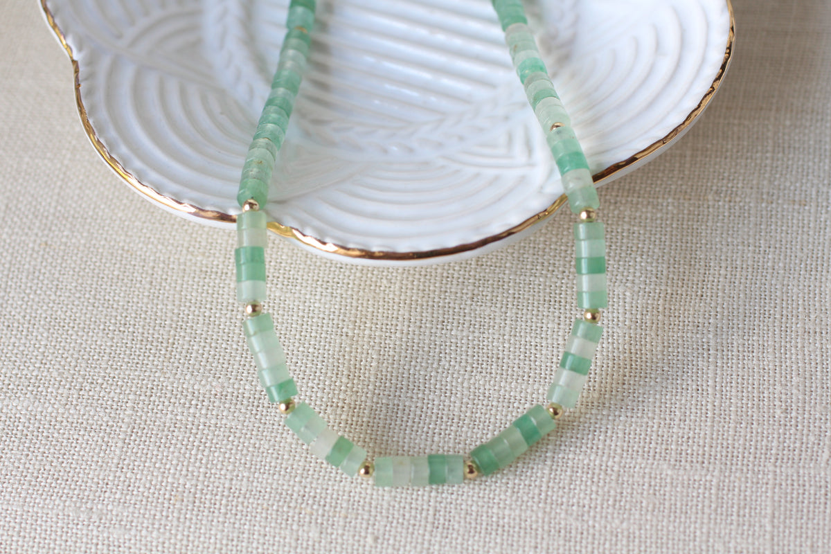A beaded necklace made of light green translucent tire shaped beads separated by small gold spacer beads, draped over a white ceramic dish with a gold-trimmed edge on a beige fabric background.