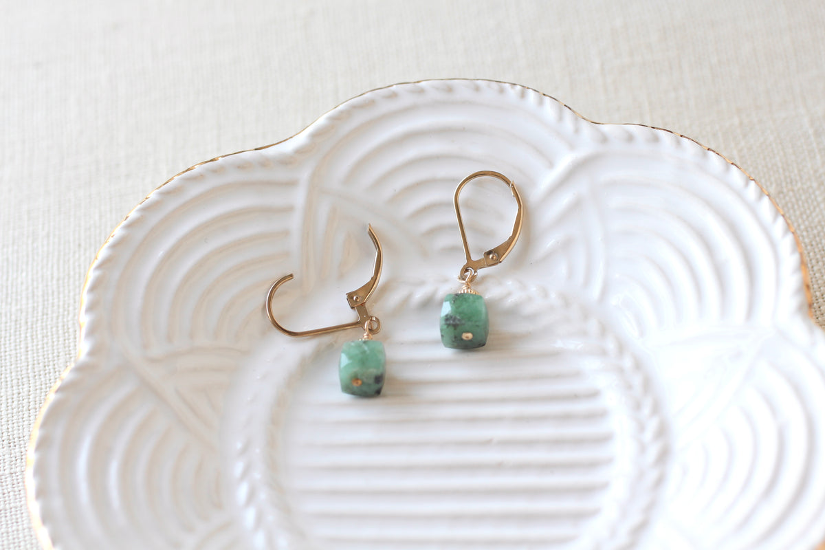 A pair of gold-tone dangle earrings with small green beads, displayed on a white scalloped ceramic dish over a light beige fabric background.