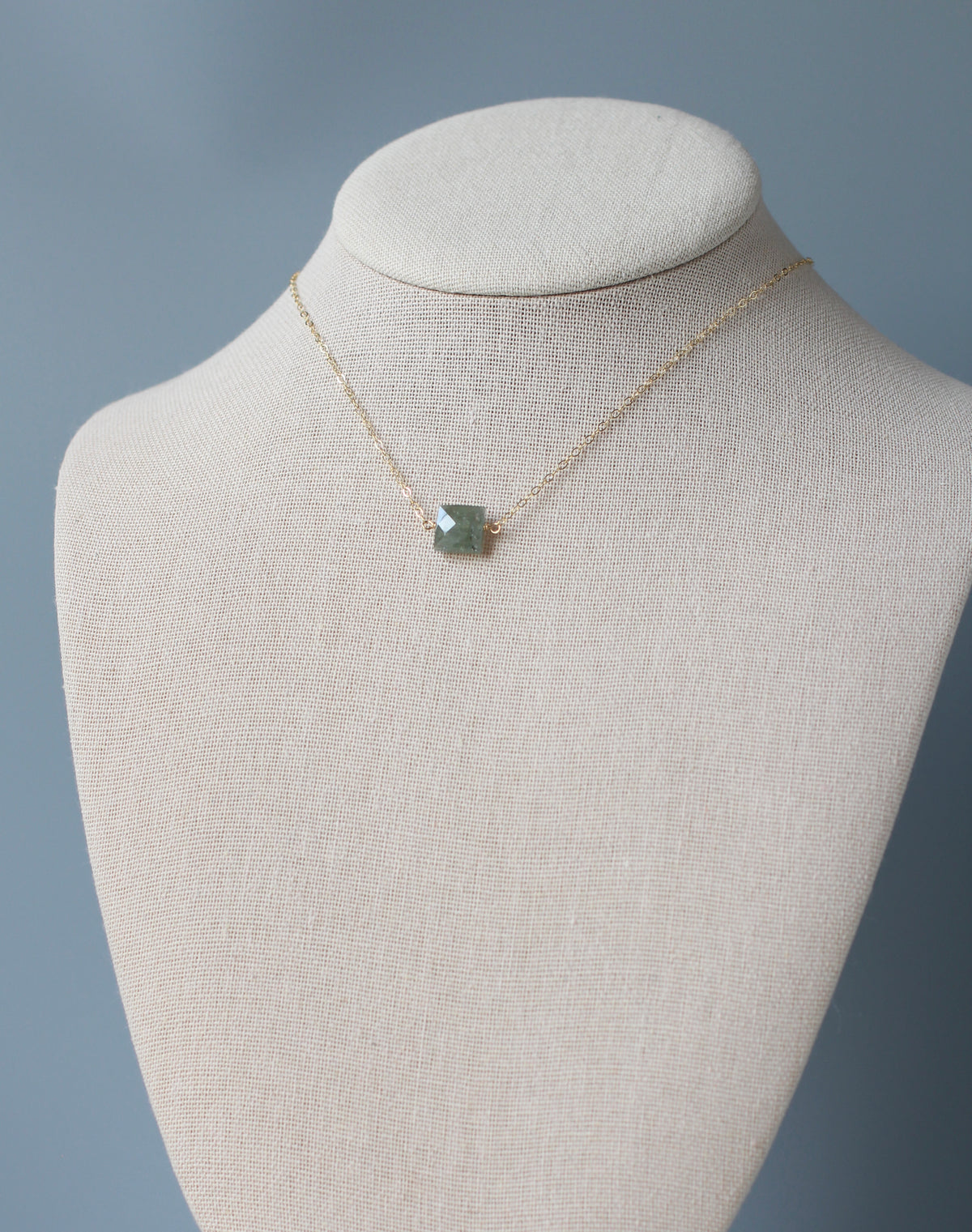 A delicate gold chain necklace with a small, square-cut green gemstone pendant, displayed on a cream-colored fabric jewelry bust against a soft blue-gray background.