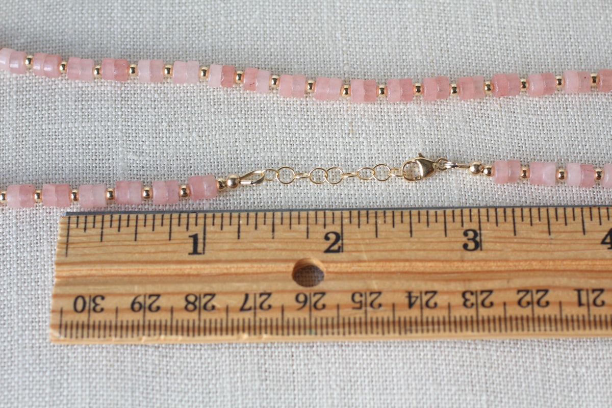 A close-up of a pink jade beaded necklace with small gold spacer beads and a gold clasp, laid on a light fabric background with a wooden ruler for scale.