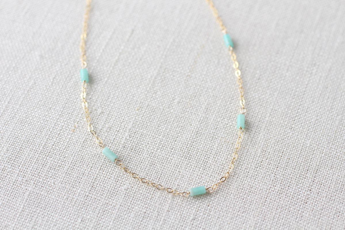 A delicate gold chain necklace featuring five small, cylindrical light blue-green beads spaced evenly along the chain, displayed on a textured off-white linen fabric background.