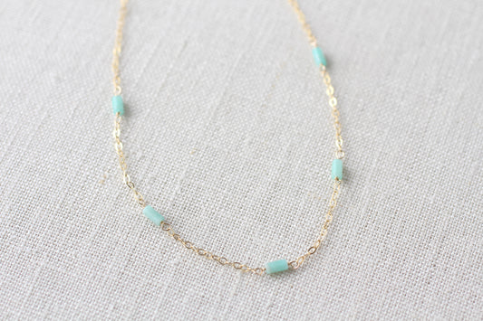A delicate gold chain necklace featuring five small, cylindrical light blue-green beads spaced evenly along the chain, displayed on a textured off-white linen fabric background.