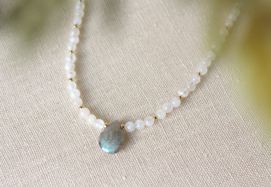 A delicate beaded necklace featuring white round beads, small gold spacer beads, and a central teardrop-shaped light blue labradorite stone pendant, displayed on a textured beige linen fabric.