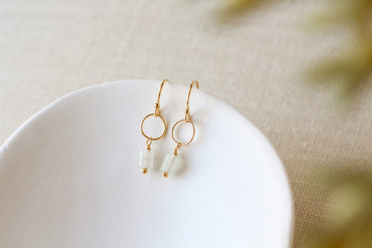 A pair of gold dangle earrings with circular hoops and small pale green bead drops, resting on a white ceramic dish against a soft beige textured background.