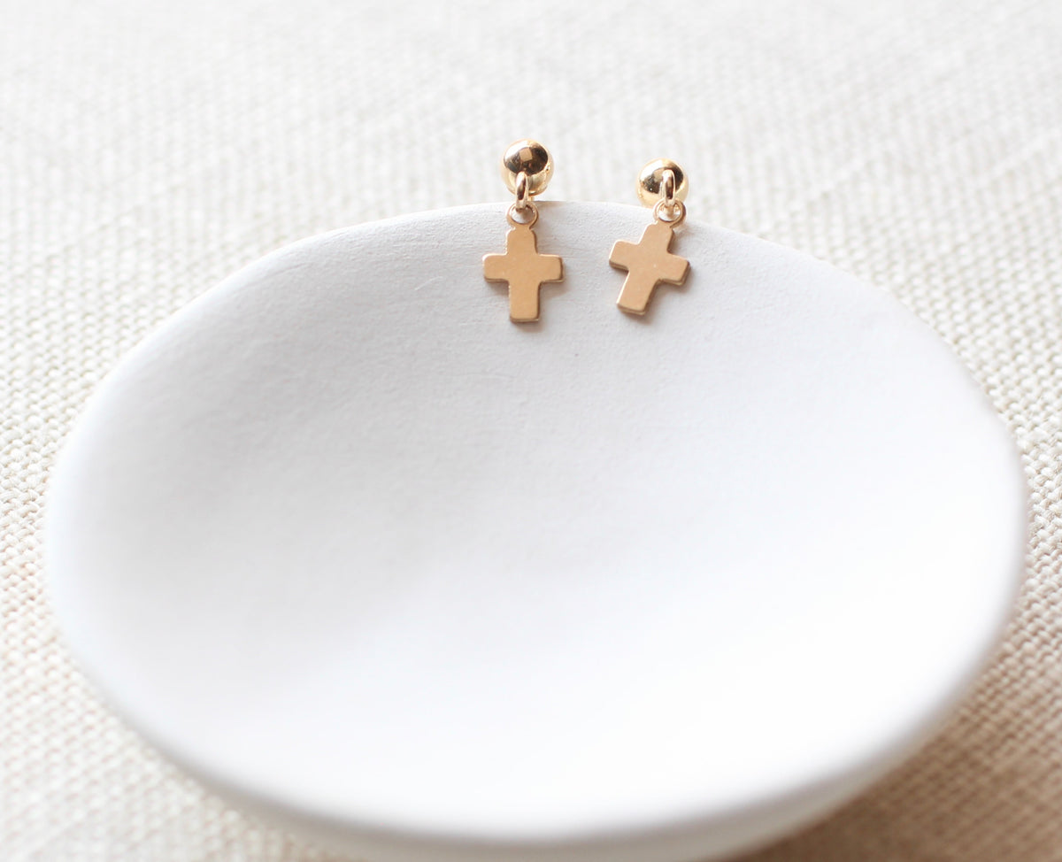 A pair of small gold cross-shaped dangle earrings displayed on a shallow white ceramic dish over a textured beige fabric background.