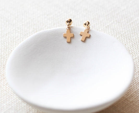 A pair of small gold cross-shaped dangle earrings displayed on a shallow white ceramic dish over a textured beige fabric background.
