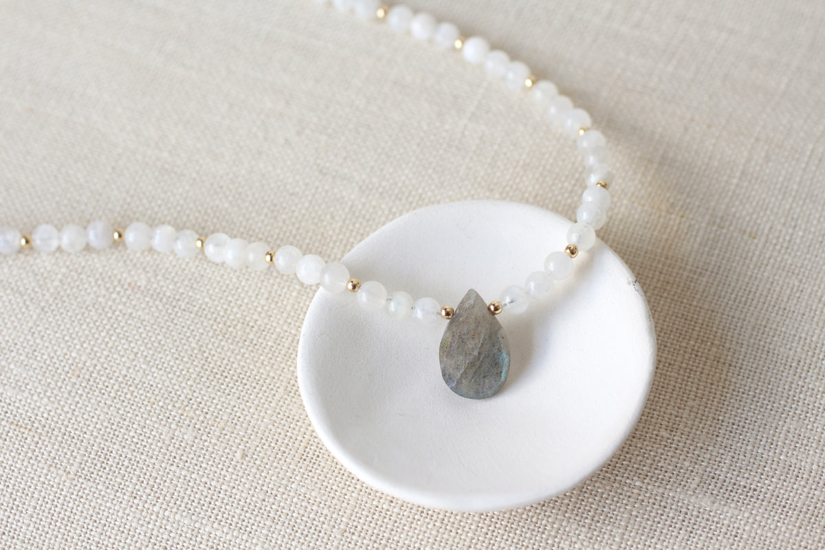 A beaded necklace with white beads and small gold spacer beads, featuring a teardrop-shaped gray labradorite pendant, displayed on a white ceramic dish over a beige linen background.
