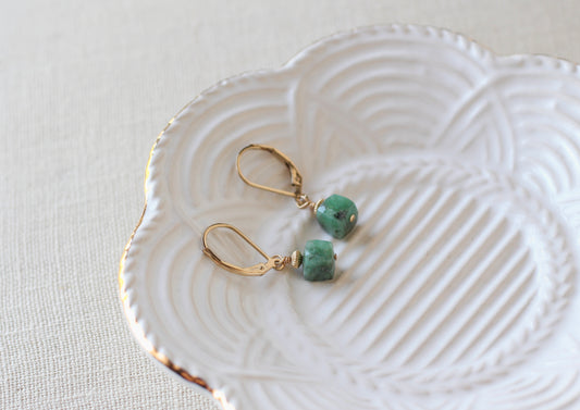 A pair of gold leverback earrings with small green stone beads, displayed on a white scalloped ceramic dish over a light beige textured fabric background.