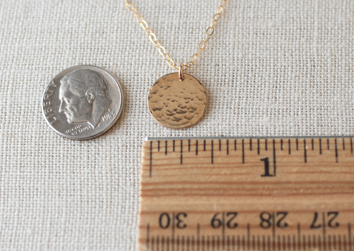 A gold pendant necklace with a textured, hammered circular disc charm is displayed next to a US dime for scale and a wooden ruler set against a neutral beige linen fabric background.