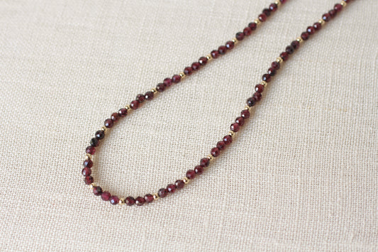 A beaded necklace made of small dark red faceted beads separated by tiny gold spacers, displayed on a textured beige linen fabric background.
