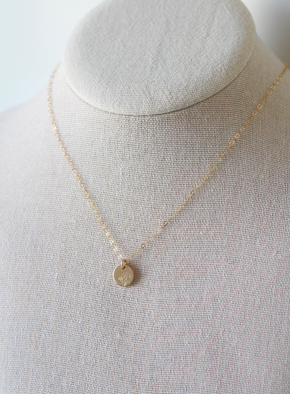 A delicate gold chain necklace with a small round pendant displayed on a textured beige fabric jewelry bust.