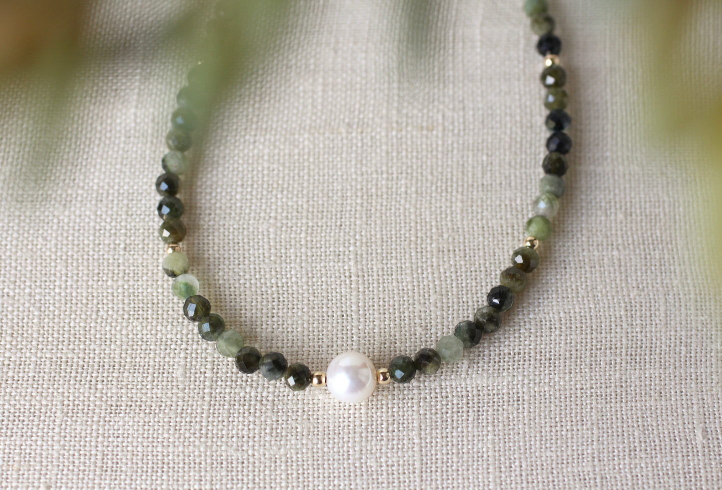 A beaded necklace featuring dark green and light green faceted beads, separated by small gold spacer rings, with a single white pearl bead at the center, displayed on a textured beige linen fabric.