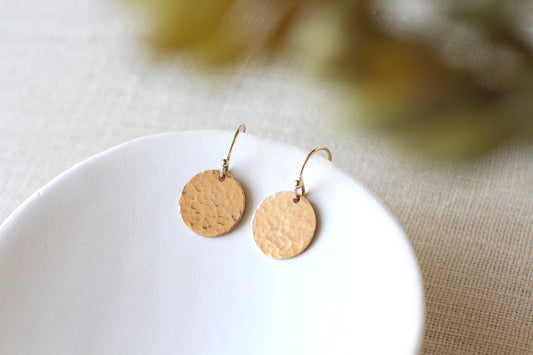 A pair of hammered round gold disc drop earrings resting on a white ceramic dish, set against a neutral beige linen background with a soft, blurred bokeh of green and yellow tones.