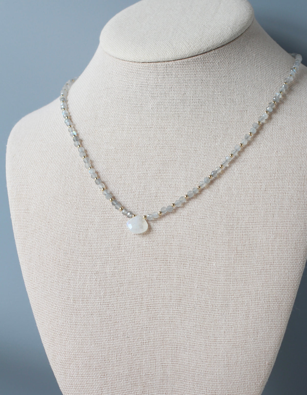 A delicate grayish labradorite beaded necklace with a white teardrop-shaped moonstone pendant, displayed on a beige fabric jewelry bust against a soft gray background.