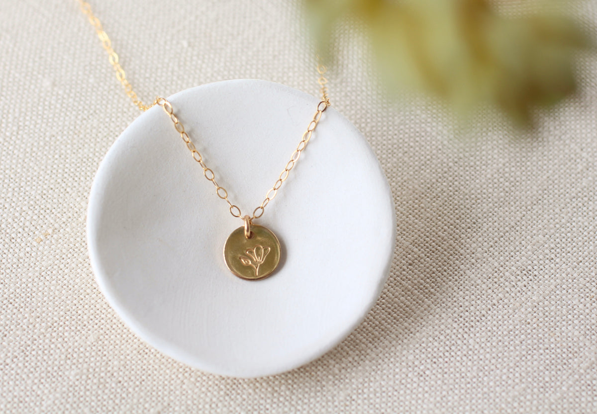 A delicate gold chain necklace with a small round gold coin pendant featuring an engraved leaf, displayed on a white ceramic dish over a beige linen background.