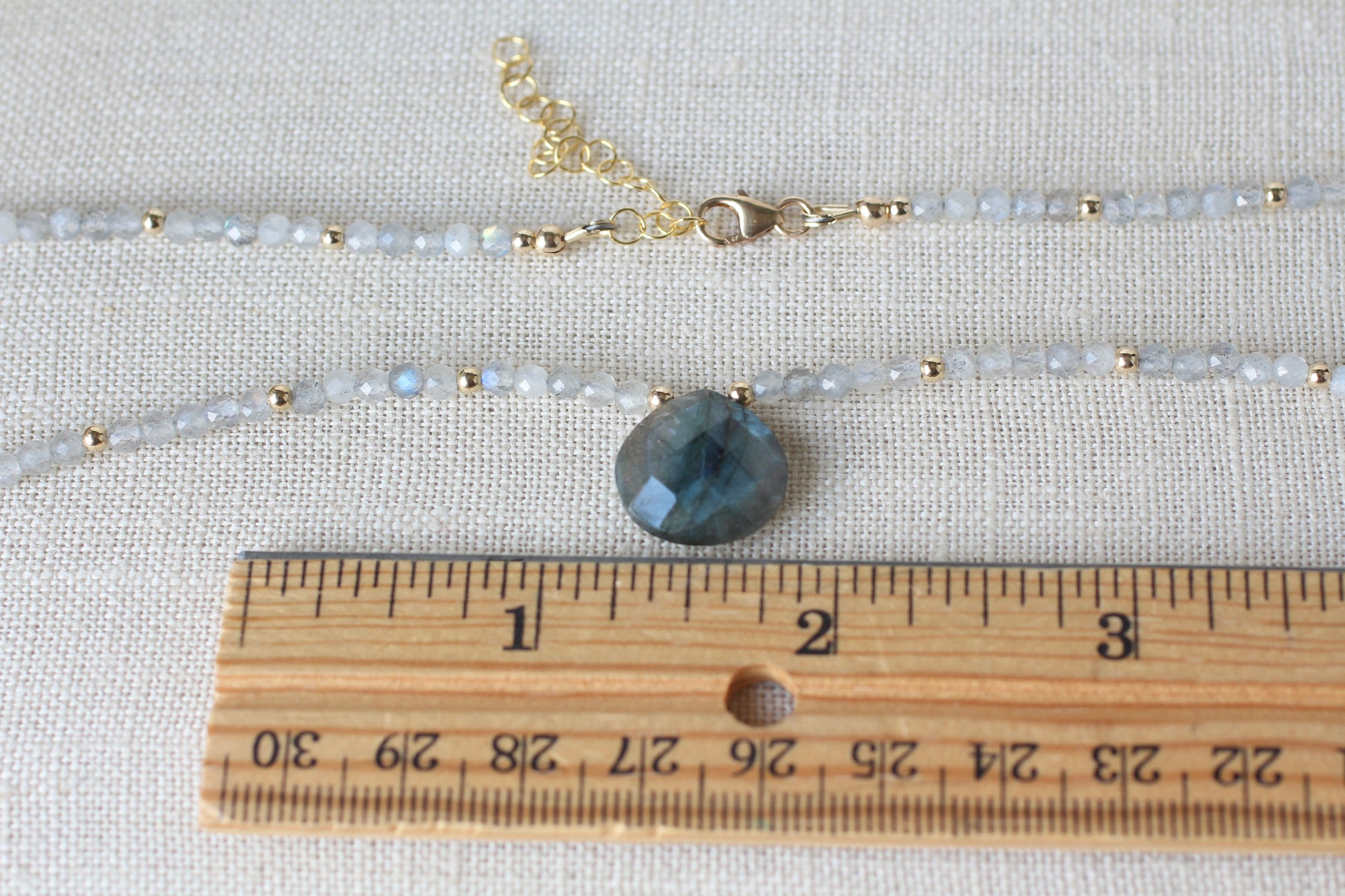 A beaded necklace with a faceted dark blue-green stone pendant, displayed on a textured fabric background with a wooden ruler for scale.