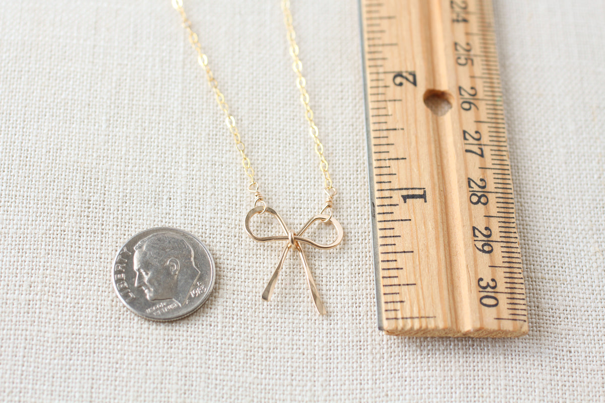 A gold necklace with a delicate bow pendant is displayed on a textured off-white fabric background, shown alongside a wooden ruler and a coin for scale.