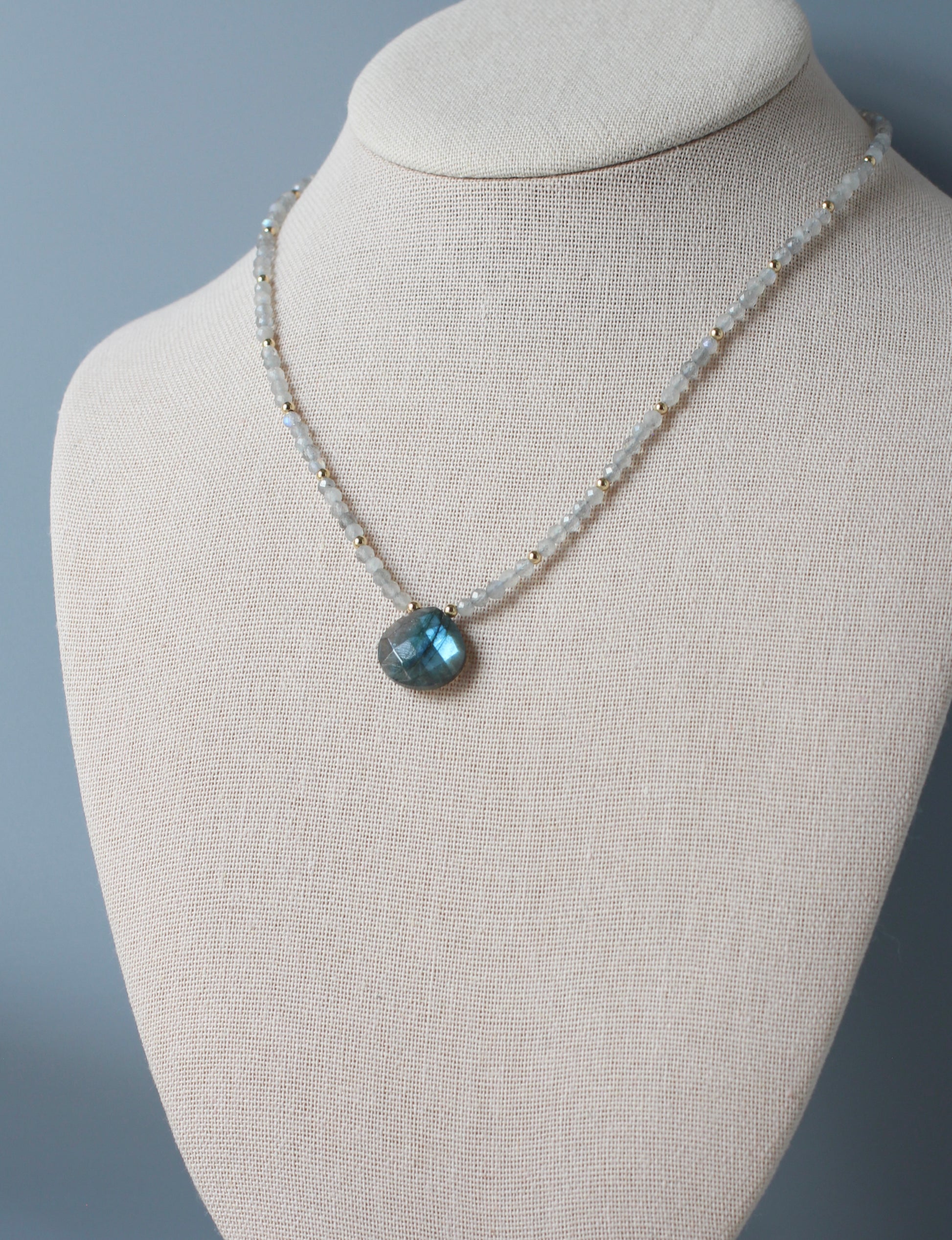 A delicate beaded necklace with a faceted blue pendant, displayed on a textured beige fabric jewelry bust against a soft gray background.