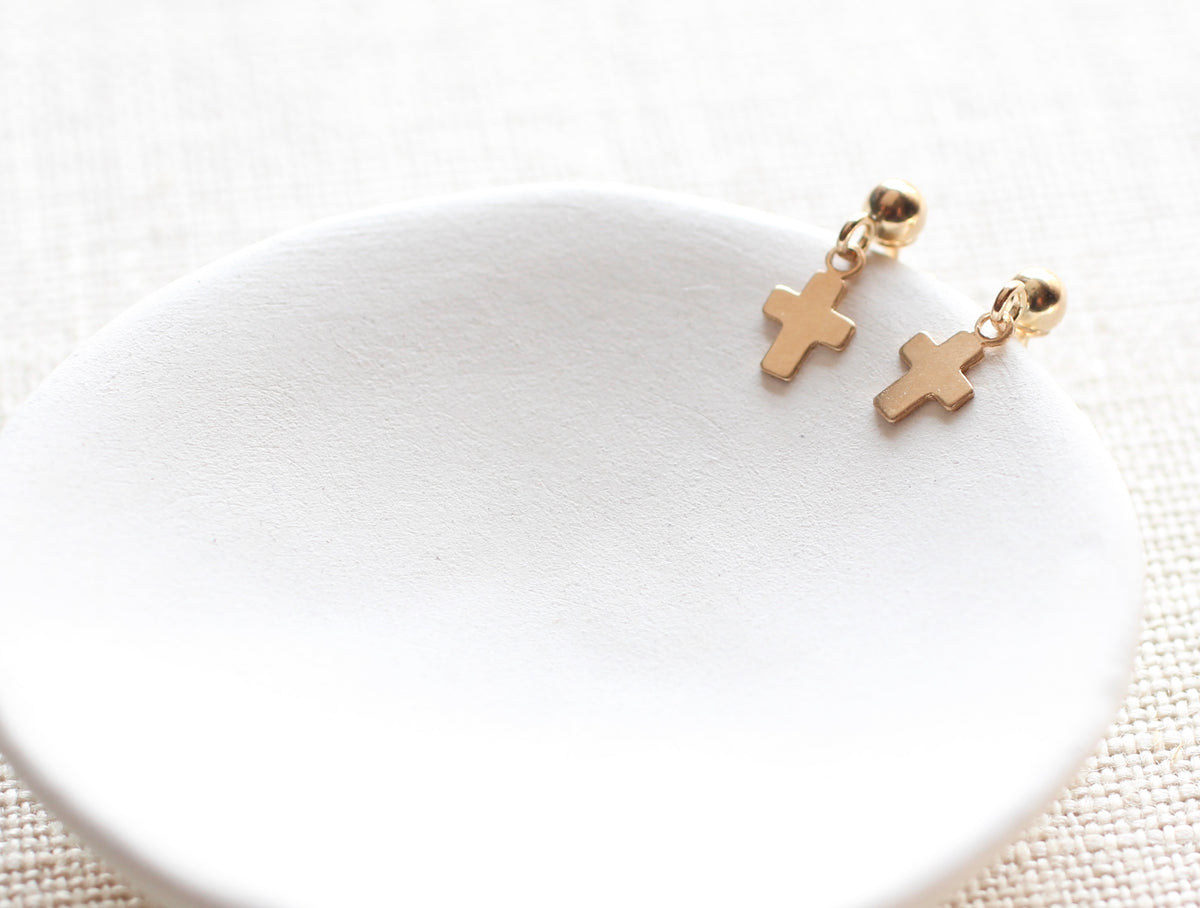 A pair of small gold cross dangle earrings resting on a white ceramic dish against a light beige textured fabric background.