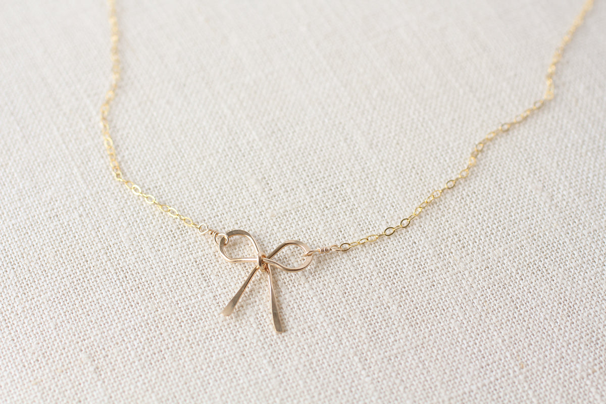 A delicate gold chain necklace featuring a small bow-shaped gold pendant, displayed on a textured off-white linen fabric background.
