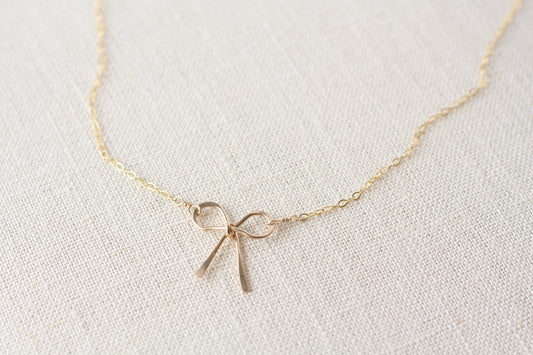 A delicate gold chain necklace featuring a small bow-shaped gold pendant, displayed on a textured off-white linen fabric background.