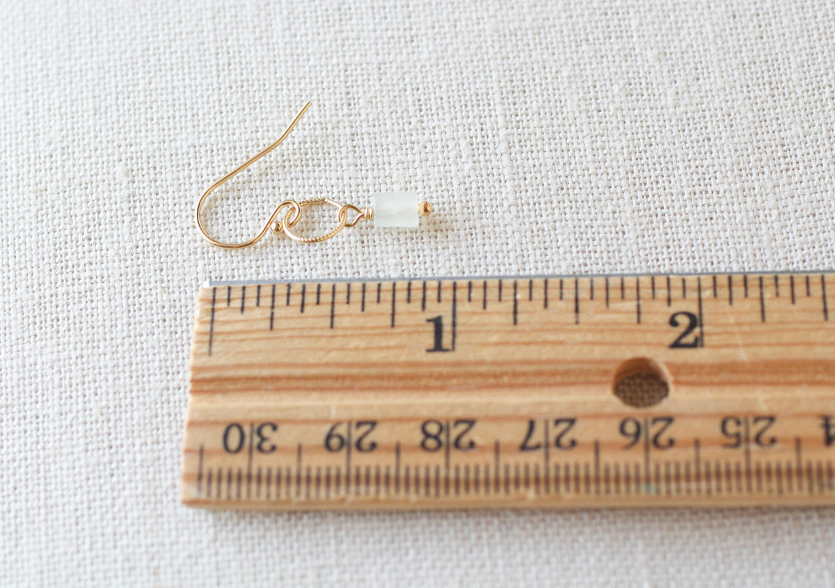 A single gold wire earring with a small pale green bead is shown next to a wooden ruler on a textured white fabric background.