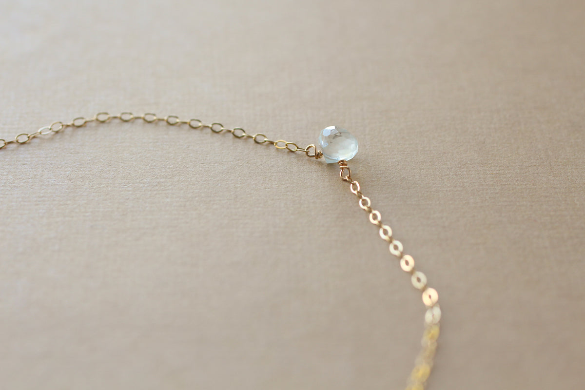 A delicate gold chain necklace featuring a small, faceted translucent light green bead, displayed on a textured beige fabric background.