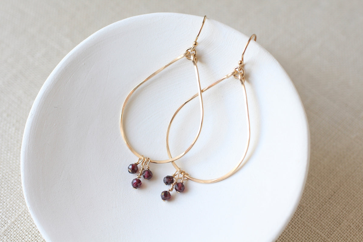 Gold teardrop hoop earrings with small dark redish purple bead clusters, displayed on a white ceramic dish over a beige linen background.