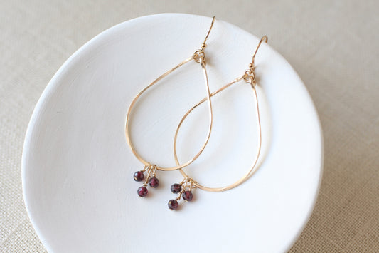 Gold teardrop hoop earrings with small dark redish purple bead clusters, displayed on a white ceramic dish over a beige linen background.