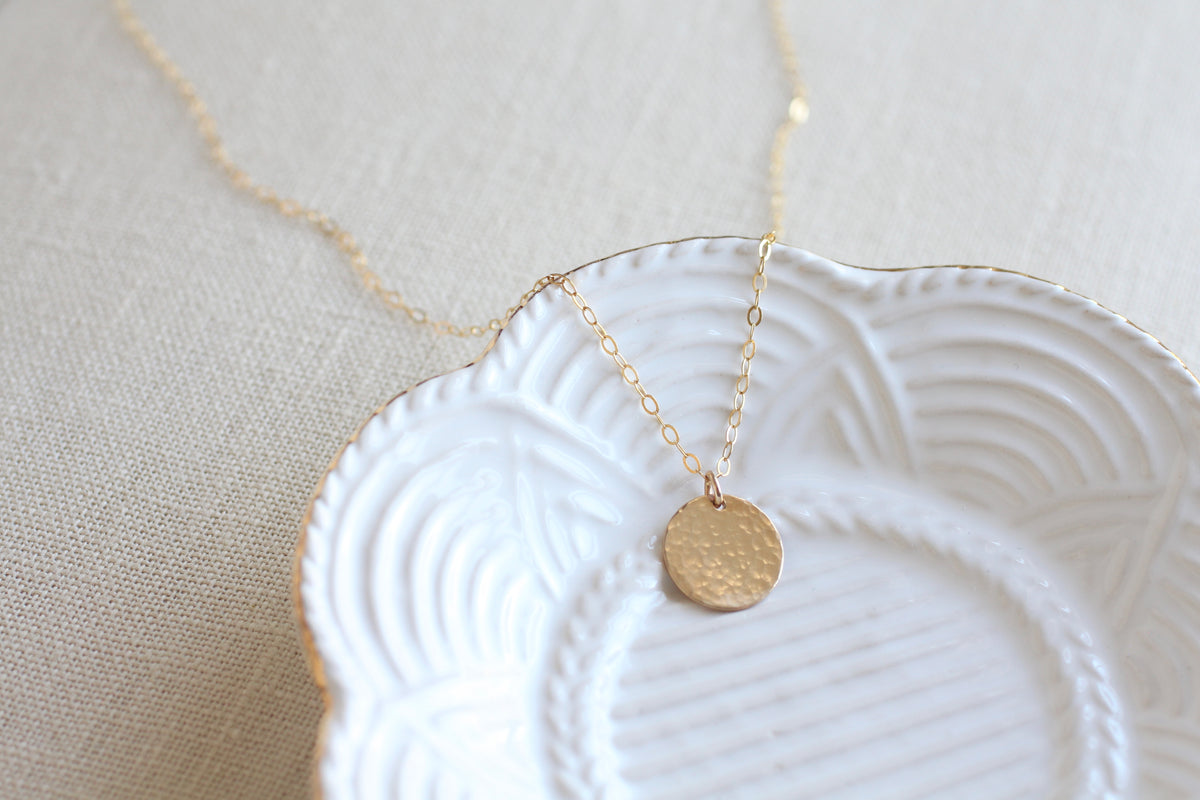 A delicate gold chain necklace with a small, textured gold disc pendant, displayed on a white scalloped ceramic dish over a beige linen background.