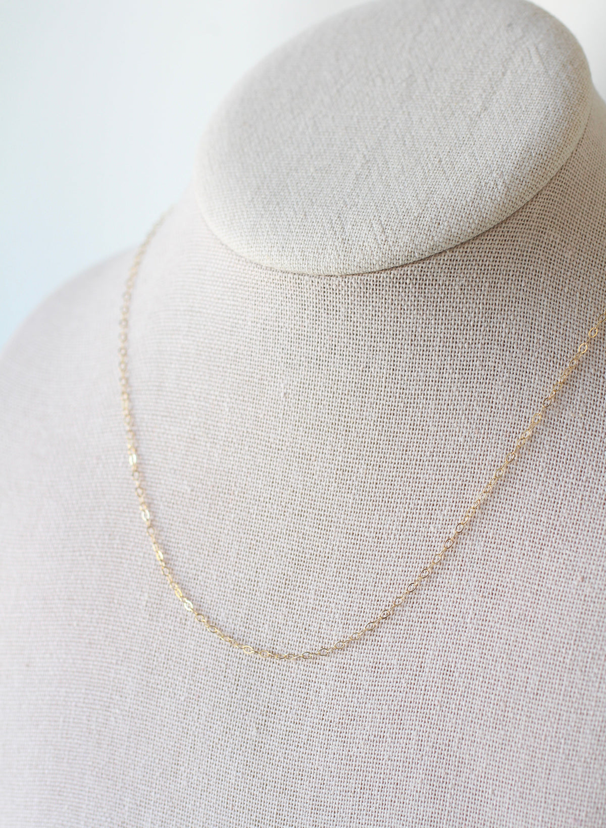 A delicate gold chain necklace draped on a textured beige fabric jewelry display bust against a soft white background.