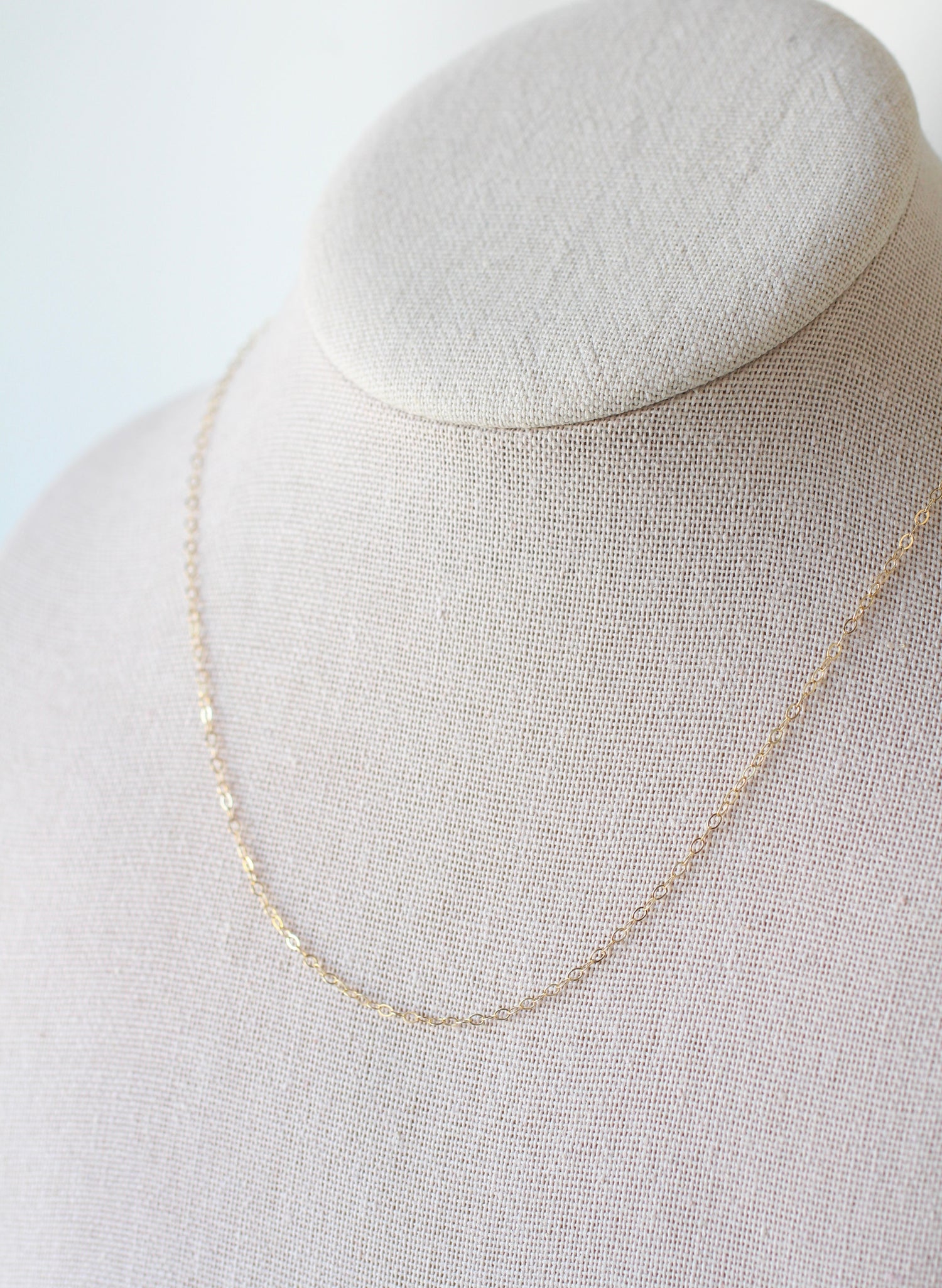 A delicate gold chain necklace draped on a textured beige fabric jewelry display bust against a soft white background.