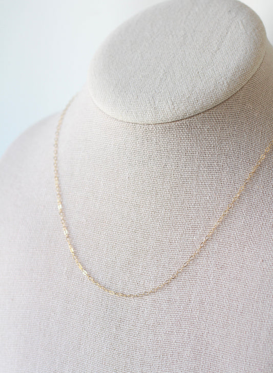 A delicate gold chain necklace draped on a textured beige fabric jewelry display bust against a soft white background.