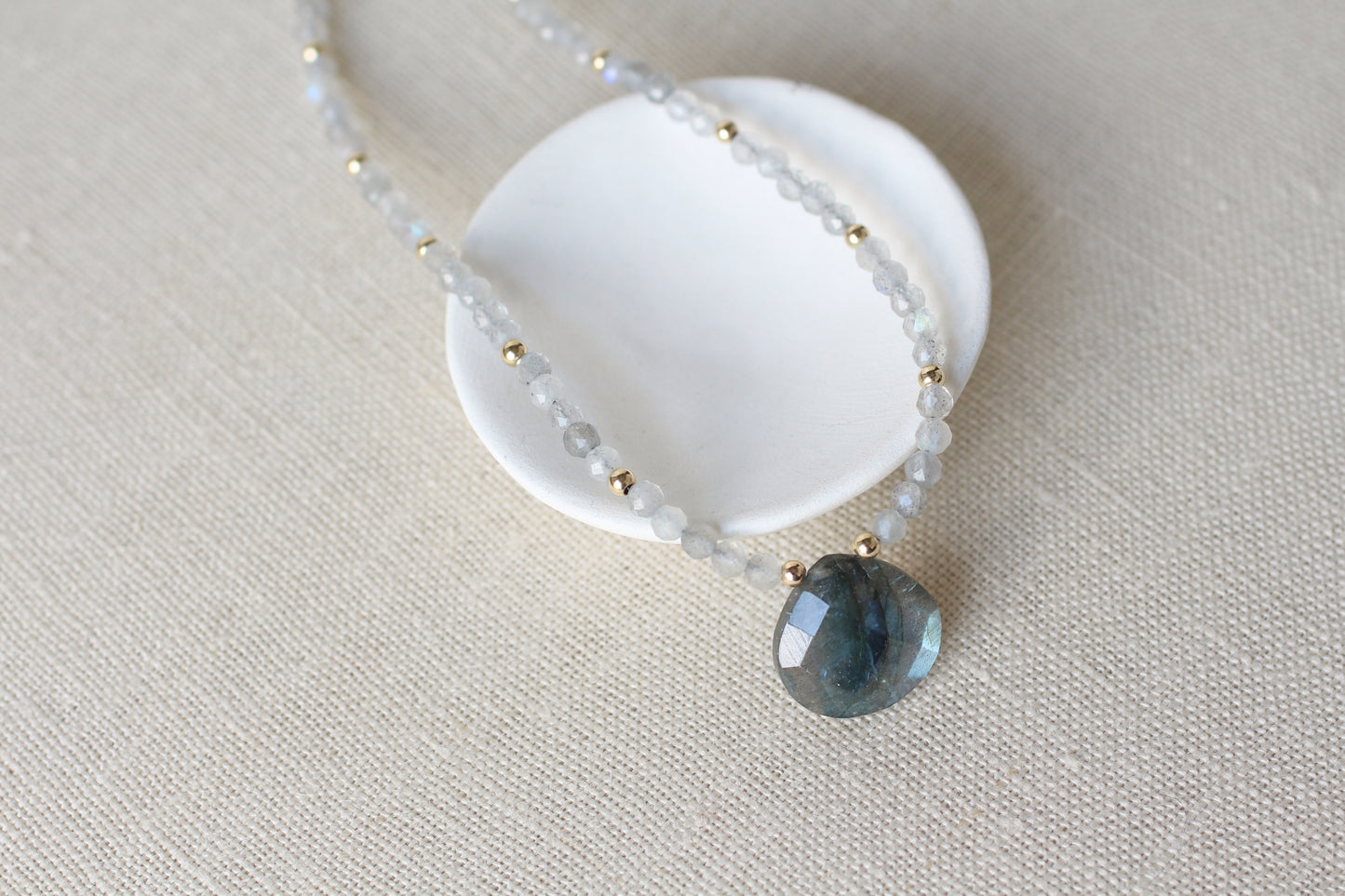 A beaded necklace with small translucent pale blue-gray beads and tiny gold spacer beads, featuring a faceted dark blue-gray stone pendant, displayed on a white ceramic stand over a beige linen background.