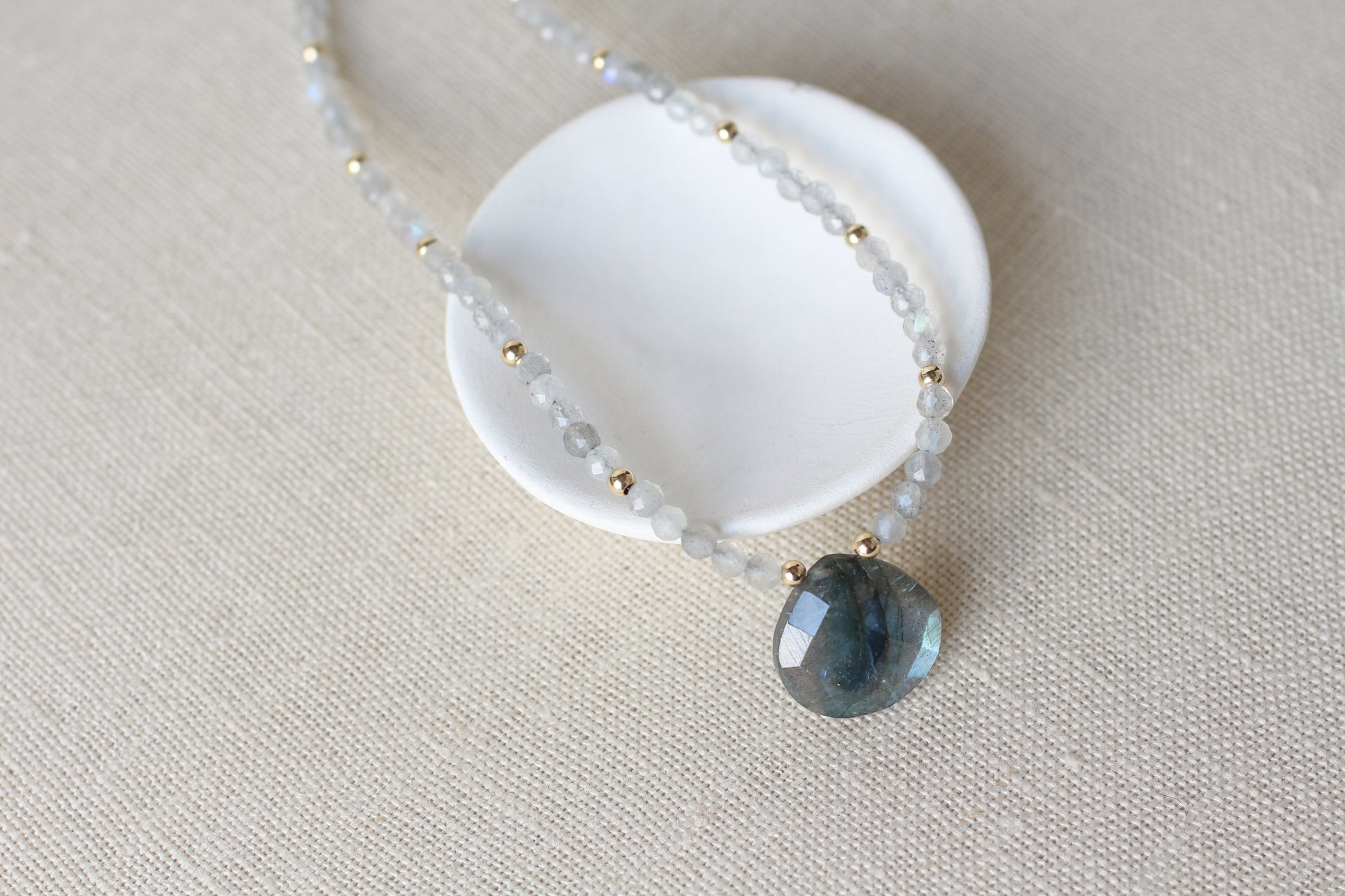 A beaded necklace with small translucent pale blue-gray beads and tiny gold spacer beads, featuring a faceted dark blue-gray stone pendant, displayed on a white ceramic stand over a beige linen background.
