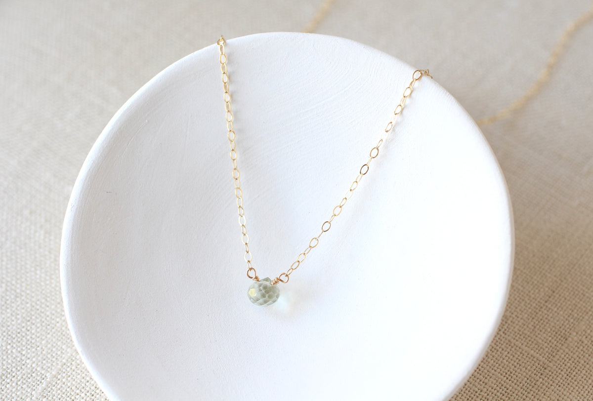 A delicate gold chain necklace with a small, faceted pale green bead pendant, displayed on a white ceramic dish over a beige linen background.