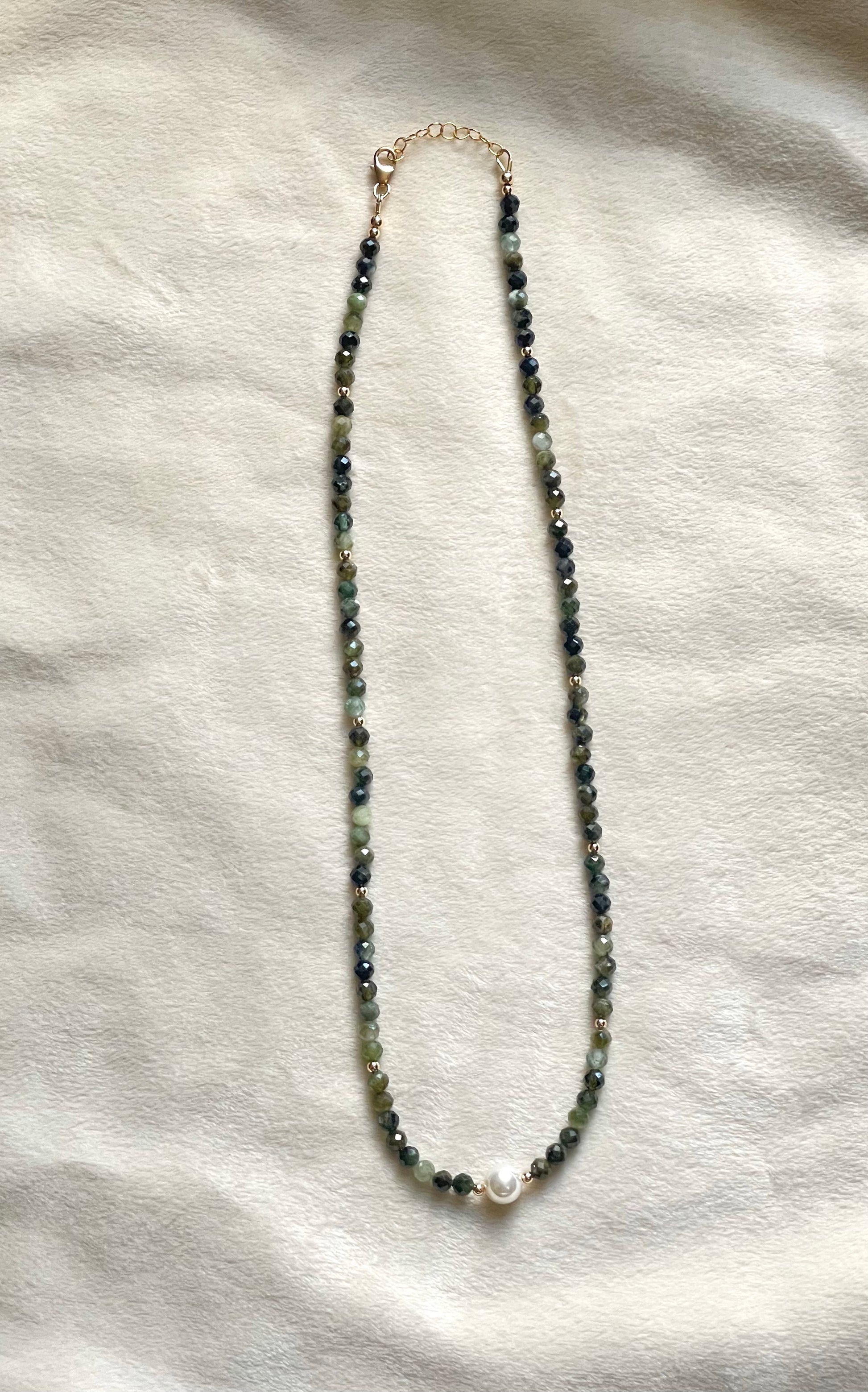 A beaded necklace with small dark green beads, a white pearl accent at the bottom center, and a gold-tone clasp, laid flat on a textured off-white fabric background.
