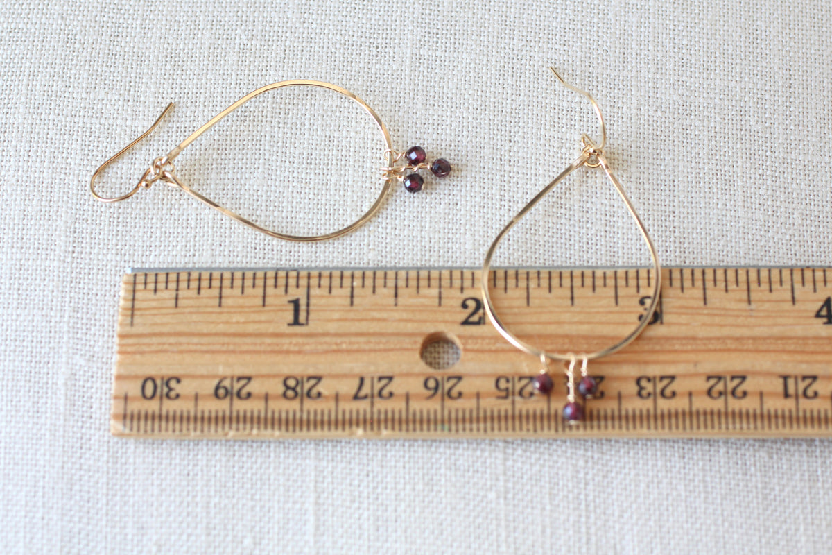 A pair of gold wire hoop earrings with small dark burgundy beads, shown next to a wooden ruler on a textured white fabric background.