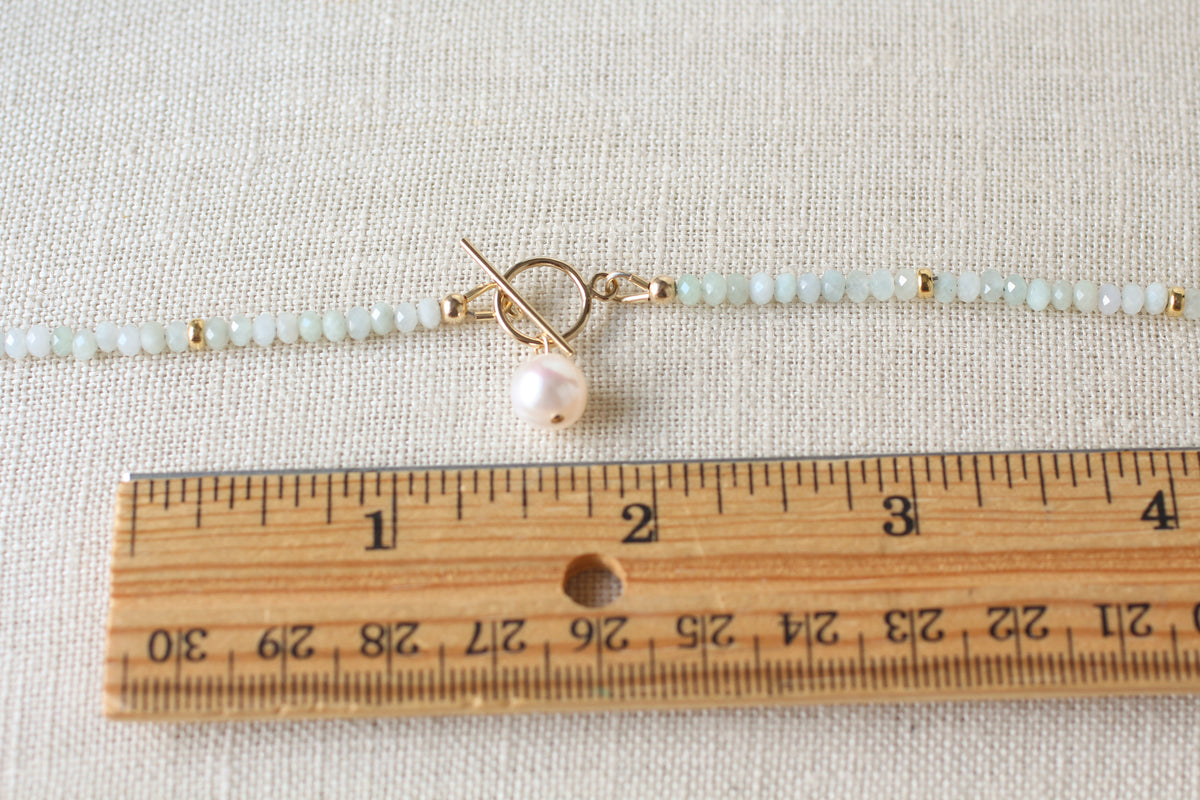 A close-up of a beaded jpale green bracelet with a toggle clasp and a pearl charm, shown next to a wooden ruler on a textured beige fabric background.