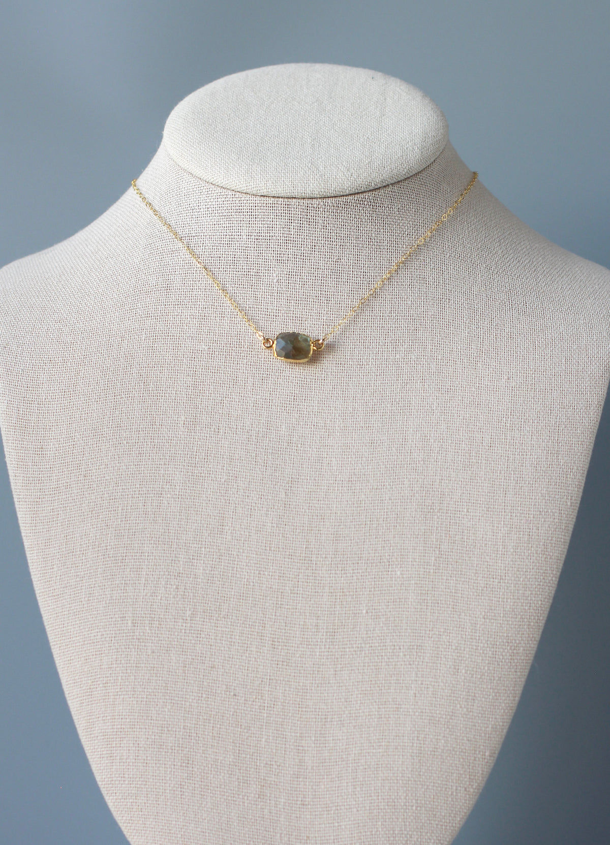 A delicate gold chain necklace with a small oval stone pendant displayed on a cream-colored fabric jewelry bust against a soft blue-gray background.