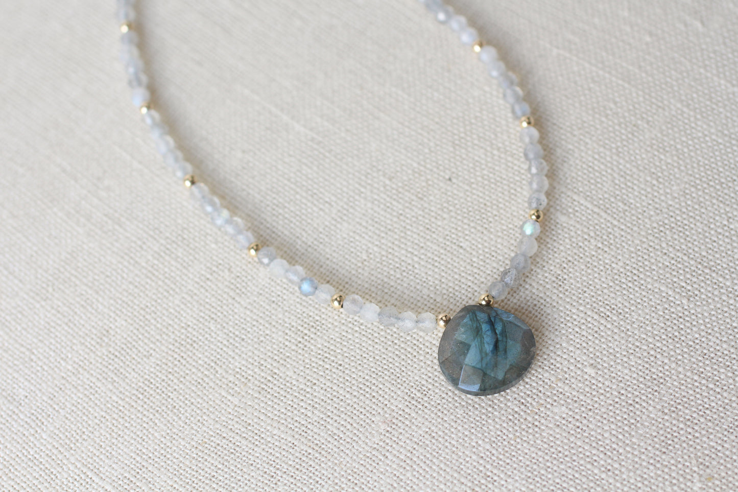 A beaded necklace featuring light blue-gray labradorite gemstone beads and small gold spacer beads, with a faceted blue-gray labradorite pendant, displayed on a textured beige linen background.