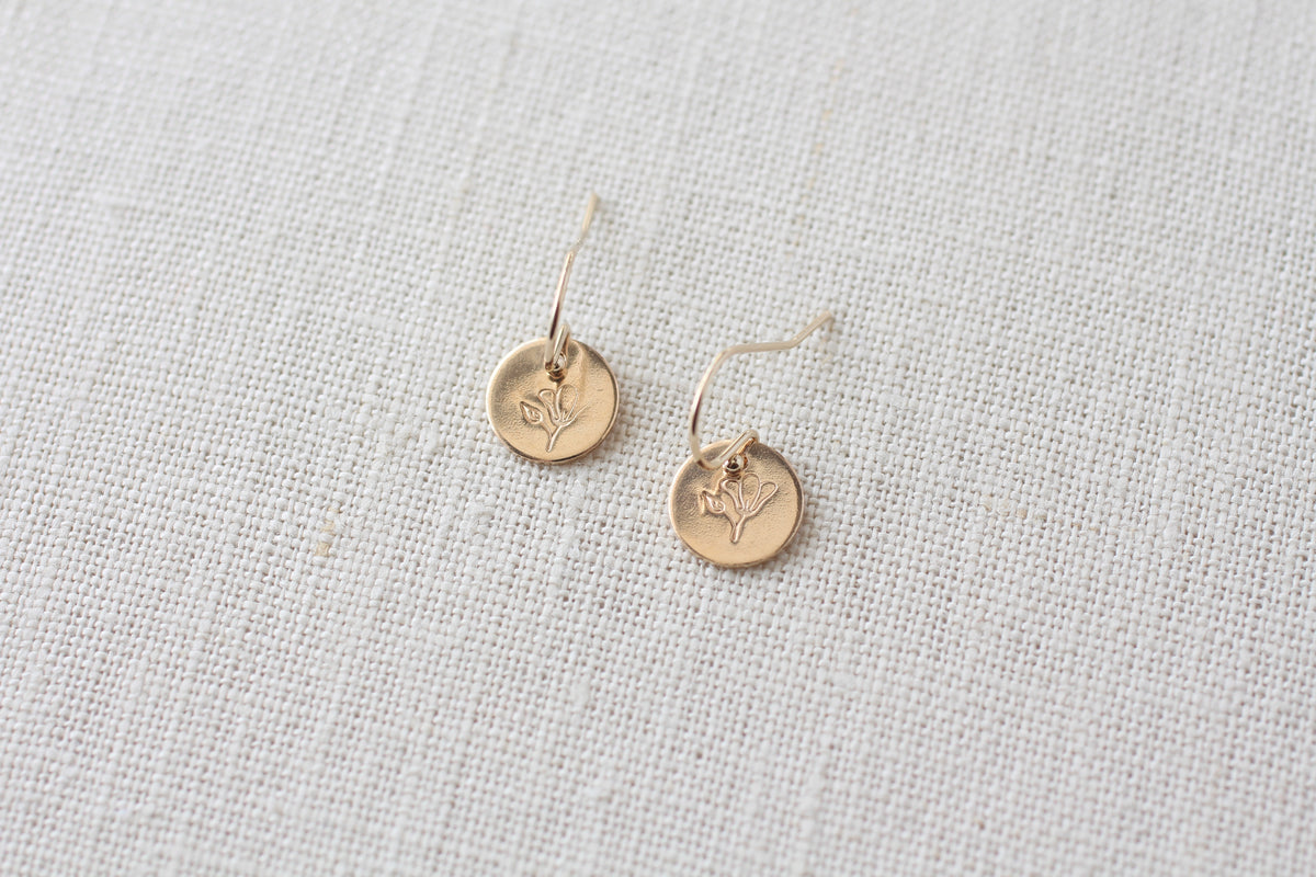 A pair of small gold disc earrings with engraved floral details, resting on a textured off-white linen fabric background.